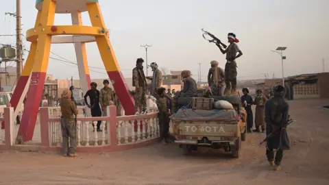 Groupe de combattants en tenue camouflage autour d'un pick-up sur une route poussiéreuse, avec un homme en silhouette tenant un fusil automatique.