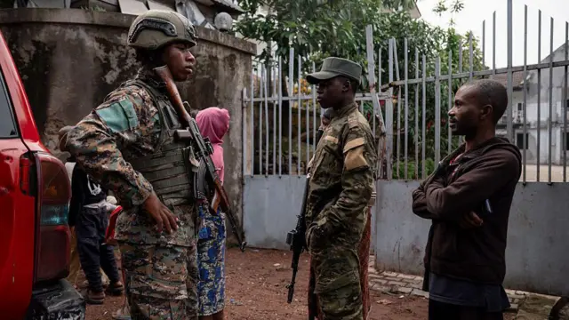 Deux militaires en uniforme et armes à la main, accompagnés d'un civil en sweat-shirt marron. Une femme en sweat-shirt rose est visible en arrière-plan.
